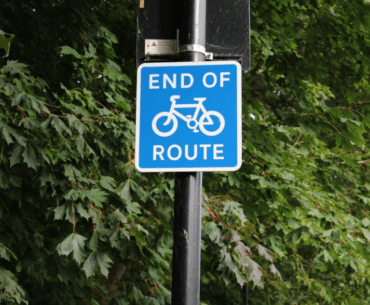 A blue and white square “End of route” sign attached to the back of a signpost. The signpost is in front of some trees, appearing to be at the side of a road.