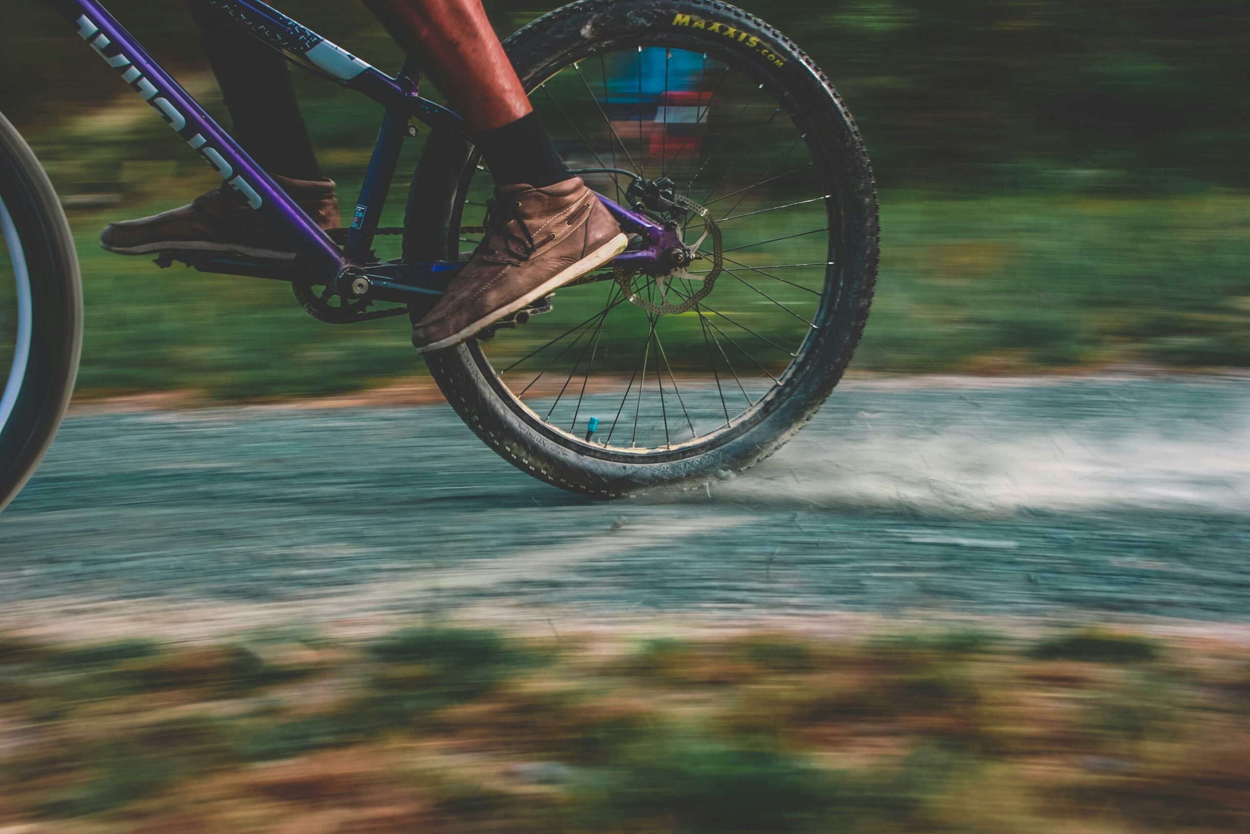 Close-up of a cyclist’s feet while they are riding a purple bicycle. The background is out of focus and shows the cyclist riding off camera towards the left.