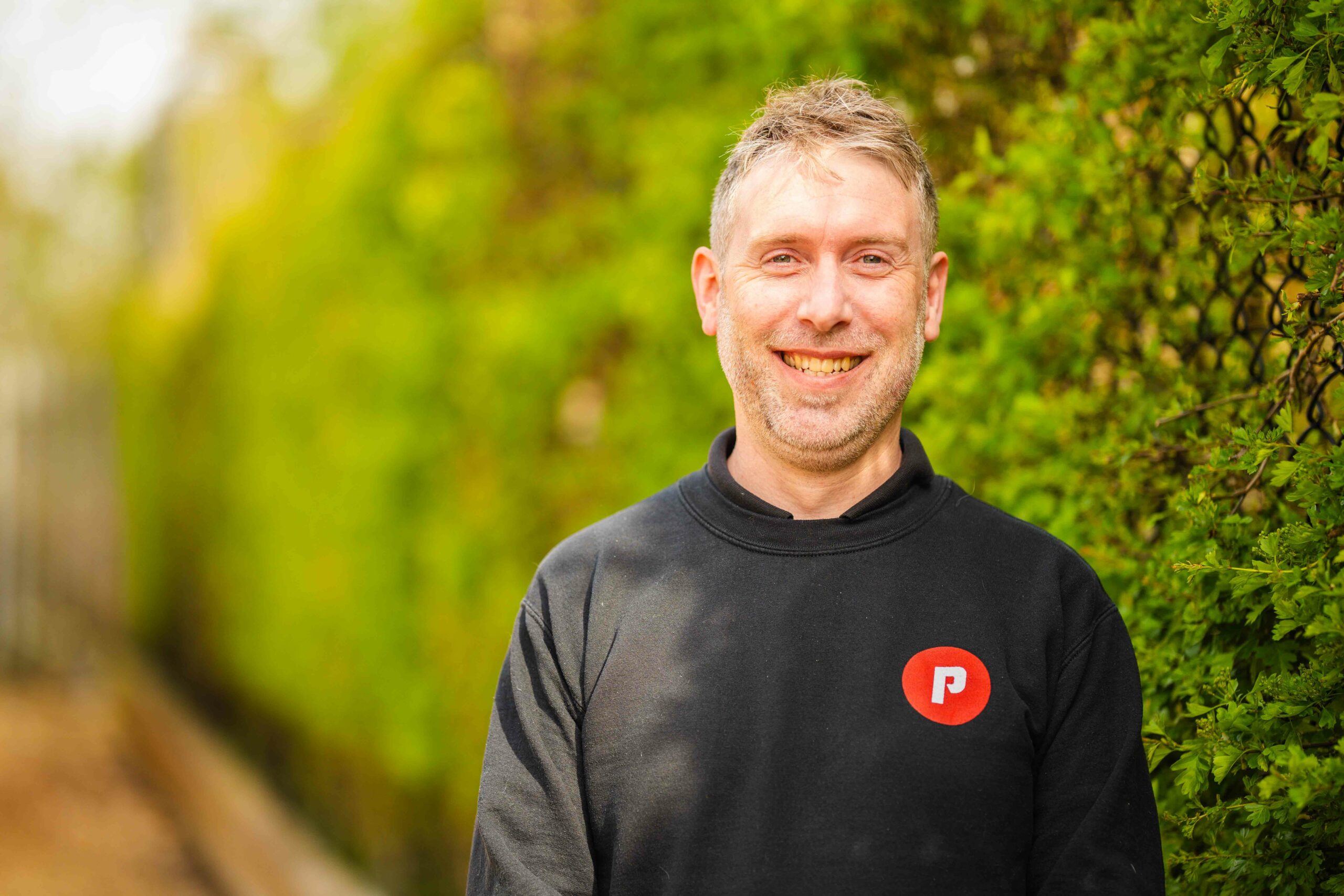 mage shows Tom Thornley, Managing Director of Paul’s Cycles, smiling and wearing a black jumper featuring Paul’s Cycles logo. He is standing in front of a hedge which is out of focus.