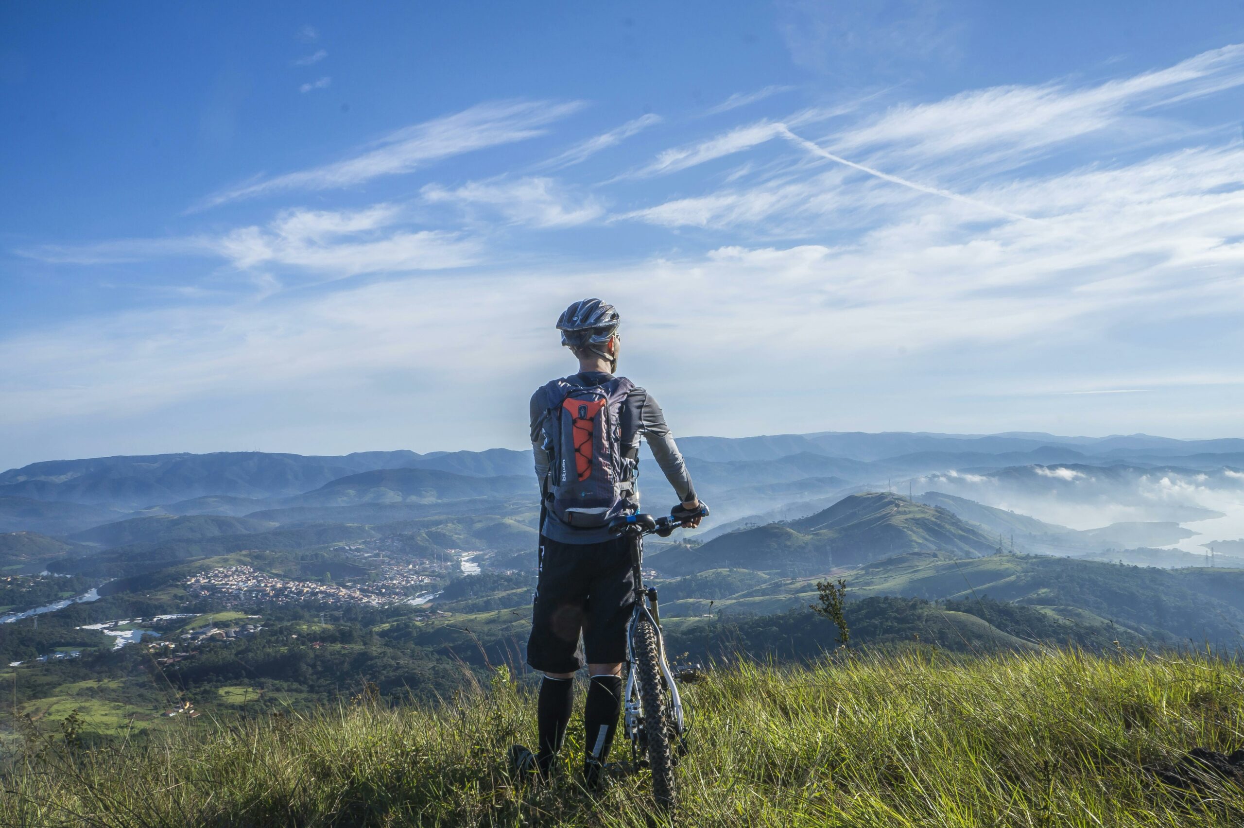 Mountain biker with a bright backpack stands on a grassy hill, overlooking a valley and mountains under a clear blue sky.
