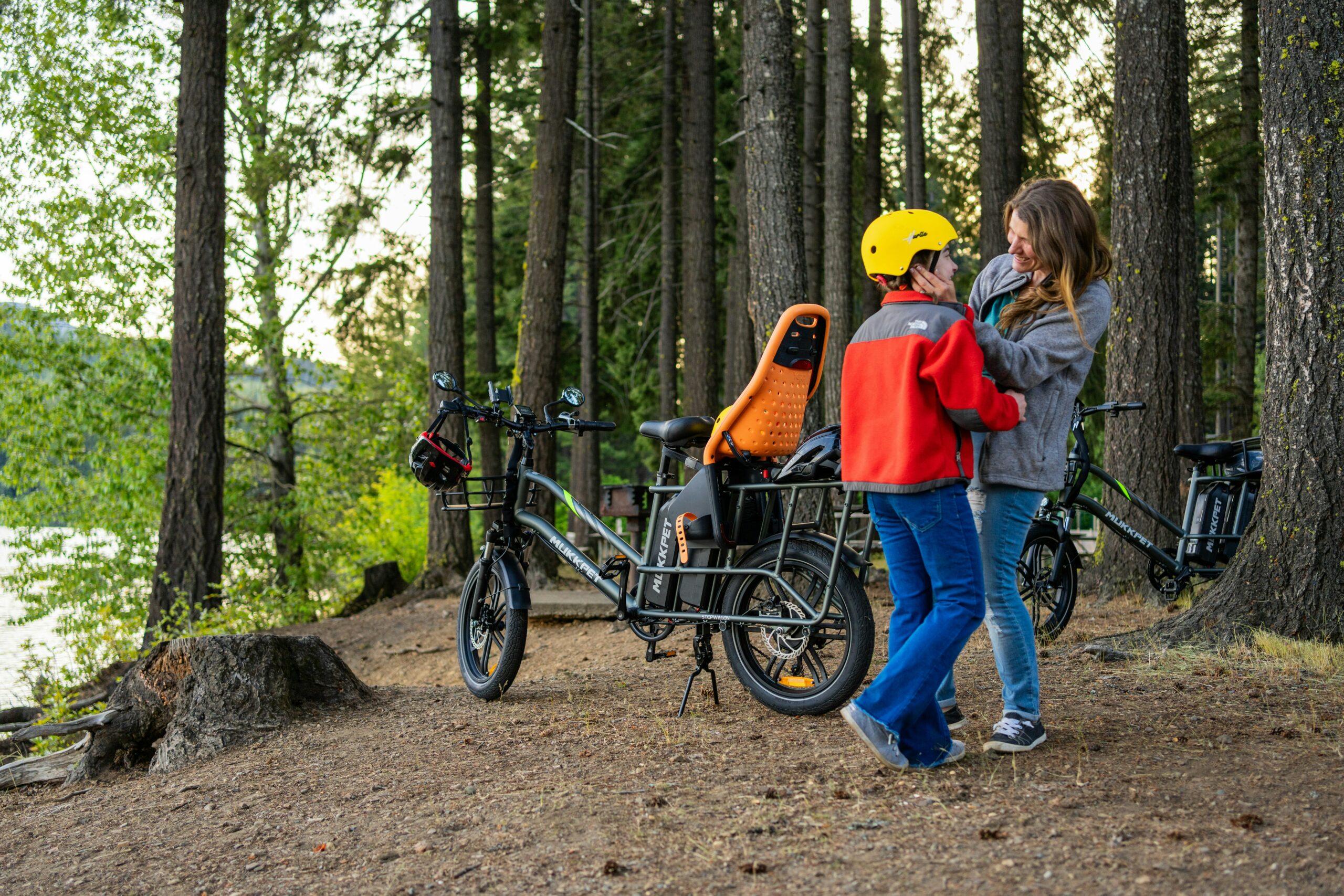 A woman adjusts a child’s yellow helmet beside two family bikes in a wooded area, with tall trees and a lake visible in the background.