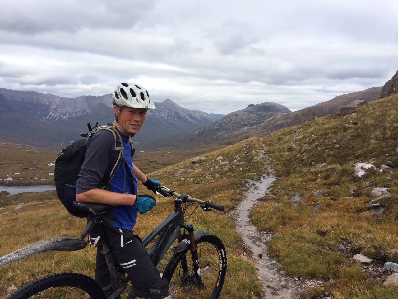Image shows James Allsopp, founder of Peak and Moor Mountain Bike Guiding, on a black bicycle in a scenic hilly terrain. He is turned to look at the camera and is wearing cycling gear.