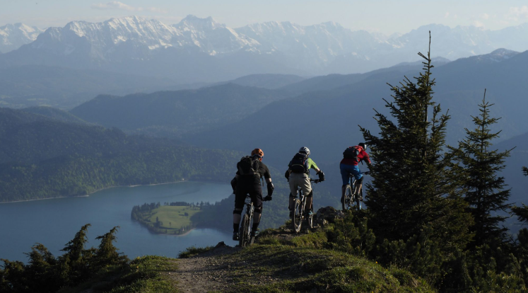 Three cyclists are cycling single file across a track on the edge of a hill. Surrounding them are trees, a lake and a scenic mountainous view.